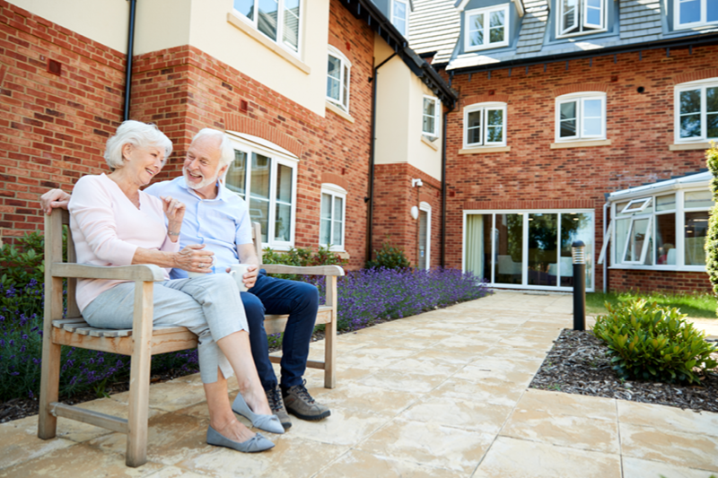 Elderly couple sitting and chatting on wooden chairs outside a care home, surrounded by gardens